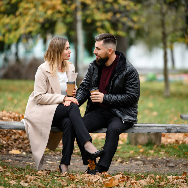 Ένα ζευγάρι, ένας άντρας και μια γυναίκα, κάθονται στο πάρκο σε ένα παγκάκι με καφέ στα χέρια τους, μια συνάντηση στο πάρκο.. - Φωτογραφία, εικόνα