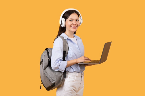 Fröhliche Studentin mit Laptop, weißen Kopfhörern und grauem Rucksack, bekleidet mit blauem Hemd, blickt in die Kamera vor gelbem Hintergrund - Foto, Bild