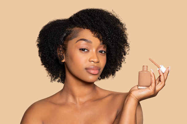 Γαλήνια Αφροαμερικανή γυναίκα με σγουρά μαλλιά που εμφανίζει μπουκάλι υγρό θεμέλιο, τονίζοντας το τέλειο ταίρι για τον τόνο του δέρματος της σε απαλό μπεζ φόντο - Φωτογραφία, εικόνα