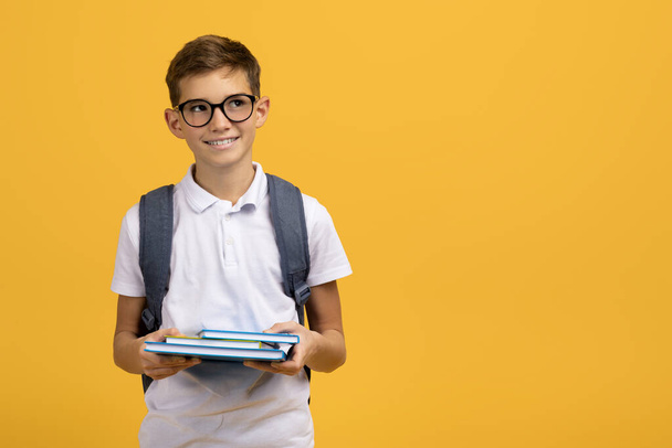 Умный школьник в очках и рюкзаке, держит учебники и смотрит в сторону на пространство для копирования, улыбающийся мальчик готов к школе, стоит на фоне желтой студии, панорама - Фото, изображение