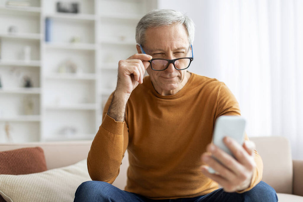 Aging eyes concept. Elderly retired grey-aired man grandfather adjusting his eyeglasses while using smartphone at home, sitting on couch, reading news, scrolling, websurfing. Vision care for seniors - Photo, Image