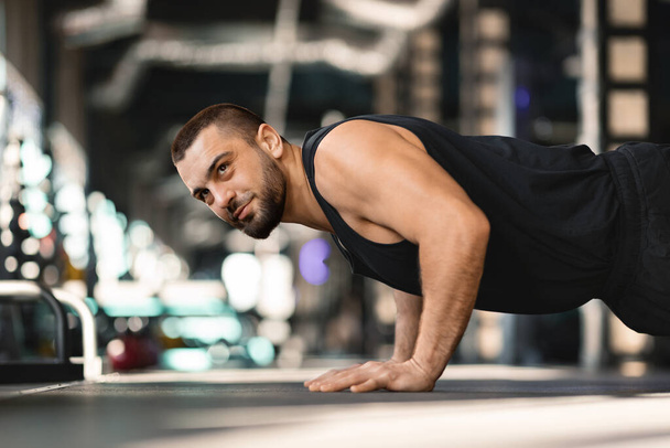 Szakállas férfi tartja deszka pozíció edzés közben a tornaterem, jóképű fiatal férfi sportoló bemutatja a mag erejét és a fókusz alatt a fitness rutin a modern sport klub, oldalnézet - Fotó, kép