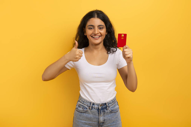 Pozitivní hezká mladá studentka z východu, ukazuje červenou plastovou kreditní kartu a palec nahoru, usmívá se na kameru, doporučuje bezkontaktní snadnou platbu, virtuální bankovnictví, izolované na žlutém pozadí - Fotografie, Obrázek