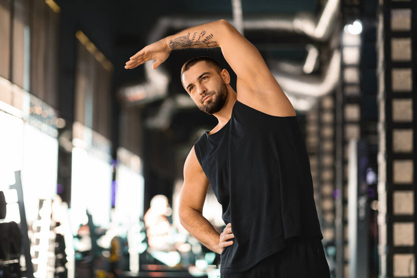 Handsome muscular man stretching his muscles before workout at modern gym, motivated male athlete bending with hands up, training in sport club interior, enjoying fitness lifestyle, copy space - Photo, Image