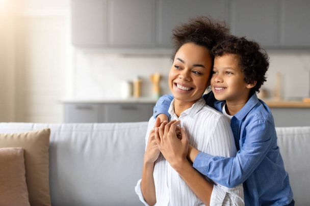 Radiant black mother and her son with curly hair share joyful hug, expressing love and happiness in their comfortable and stylish home, looking at free space - Photo, Image