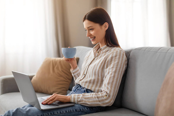 Sonriente mujer joven relajada disfrutando de una taza de café mientras trabaja casualmente en su computadora portátil o navegar por Internet, sentado en un sofá gris, espacio libre - Foto, imagen