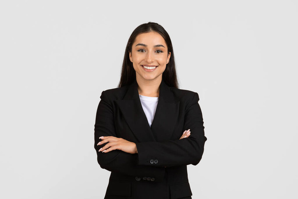Jovem empresária europeia profissional com sorriso radiante, vestida com um elegante terno preto, segura com os braços cruzados contra fundo cinzento - Foto, Imagem