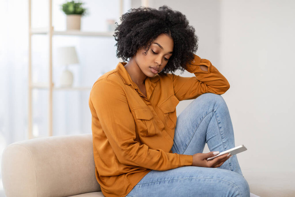 Pensive surullinen Afrikkalainen Amerikkalainen nainen kihara tukka, yllään sinappi pusero, istuu sohvalla katsomassa hänen puhelimensa mietiskelevä ilme, jutteleminen, lukea uutisia - Valokuva, kuva