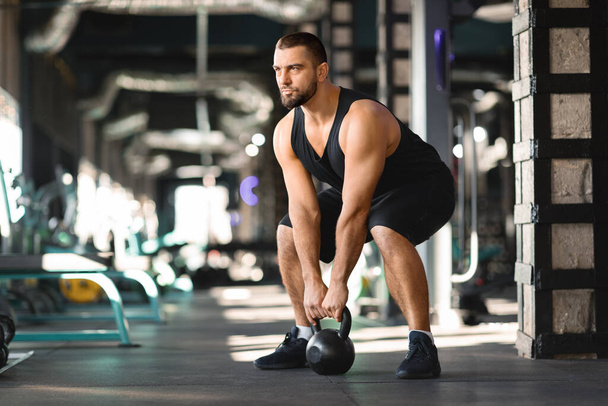 Motivated Muscular Man Training With Kettlebell At Modern Gym Interior, Young Handsome Bodybuilder Lifting Heavy Dumbbell While Exercising In Sport Club, Training Muscles, Copy Space - Photo, Image