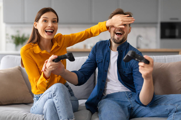 Jóvenes cónyuges alegres dedicados a jugar videojuegos, con la mujer riéndose cubriendo los ojos del hombre, creando un ambiente hogareño juguetón, sentados en el sofá - Foto, imagen