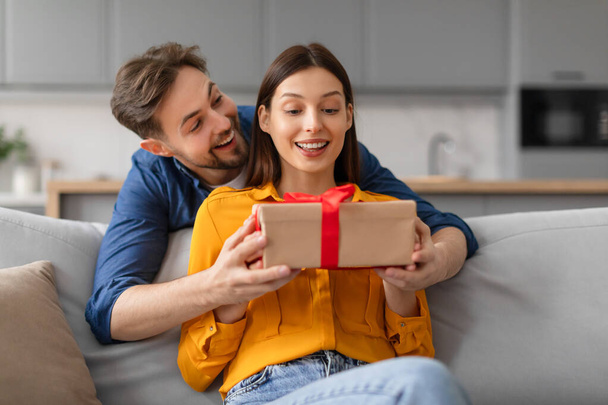 Jeune femme excitée recevant un cadeau surprise de l'homme debout de dos dame assise sur le canapé, souriant et capturant moment de joie et d'affection - Photo, image