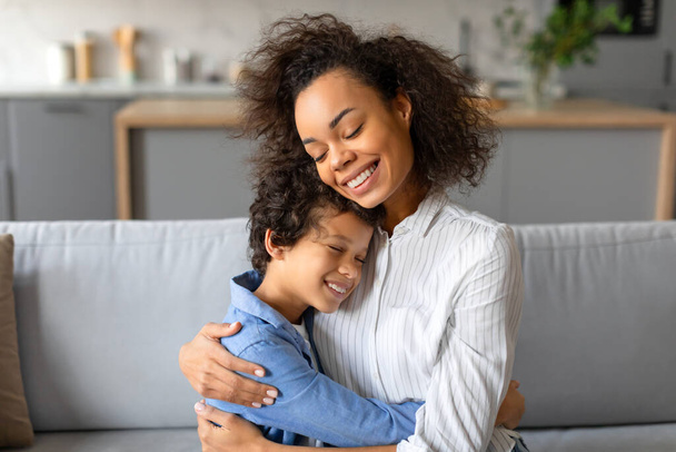 Mère noire affectueuse étreignant son fils préadolescent, souriant avec bonheur et chaleur, dans un cadre lumineux et confortable, assise sur un canapé - Photo, image