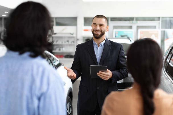 A professional car salesman in a well-tailored suit confidently discusses vehicle options with a young couple at a spacious and modern car dealership, digital tablet in hand. - Photo, Image