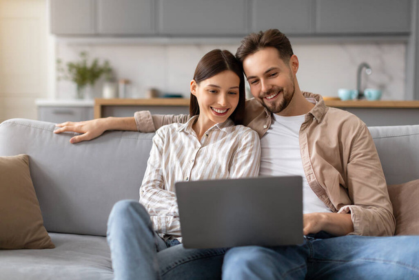 Jovens casais felizes sentados no sofá, envolvidos com o computador portátil juntos navegando na internet em um ambiente confortável em casa, irradiando alegria e união - Foto, Imagem