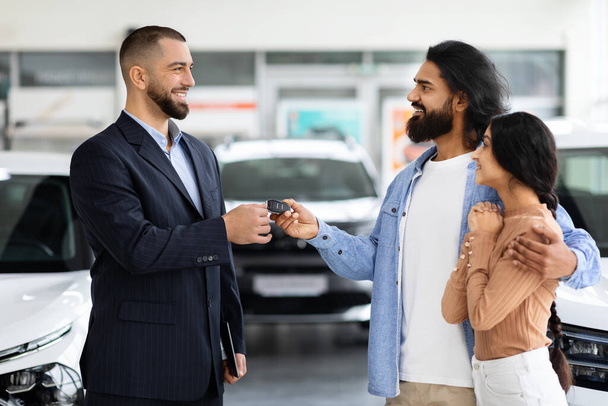 Car purchase or lease concept. Cheerful young indian couple taking car key from auto salesman at modern dealership. Millennial eastern spouses buying new automobile at showroom store - Photo, Image
