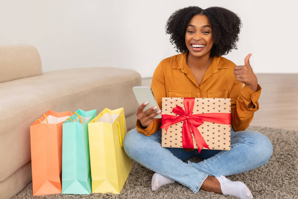 Szczęśliwego tysiąclecia Afroamerykanka z szerokim uśmiechem trzyma prezent i jej smartfon, dając kciuk w górę siedząc na podłodze w pobliżu kolorowych toreb na zakupy w domu - Zdjęcie, obraz