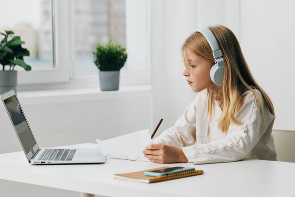 Щаслива кавказька дівчина, яка навчається вдома, використовує ноутбук для навчання та онлайн-ігор, з навушниками та ноутбуком на столі Сучасне цифрове з'єднання забезпечує віртуальний шкільний досвід, - Фото, зображення
