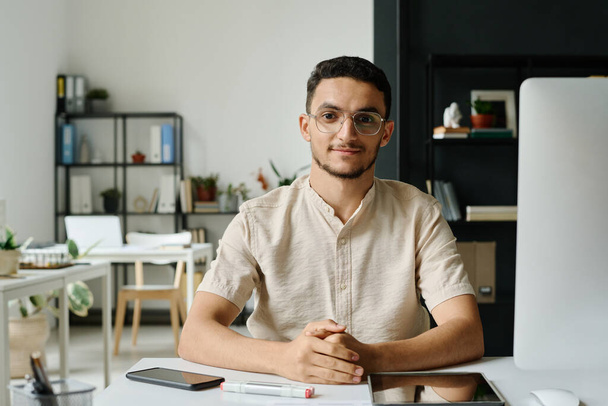Mladý muslim muž ceo nebo podnikatel sedí na pracovišti s stolním počítačem v kopracovním prostoru a dívá se do kamery - Fotografie, Obrázek