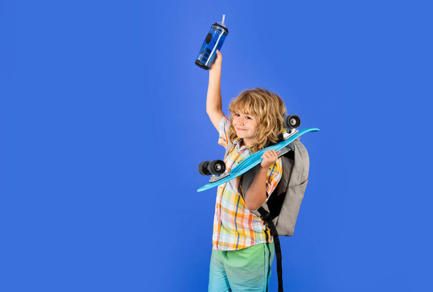 青い隔離された背景にロングボードとウォーターボトルを保持する子供の少年. ペニーボードの子供。 ペニーボード付き陽気な小さなファッションの子供のスタジオショット - 写真・画像
