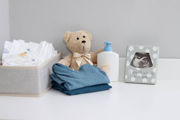 Een stapel luiers, rompertjes, teddybeer en babyspullen op witte commode, verpleegkamer  - Foto, afbeelding