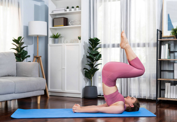 Flexible and dexterity woman in sportswear doing yoga position in meditation posture on exercising mat at home. Healthy gaiety home yoga lifestyle with peaceful mind and serenity. - Photo, Image