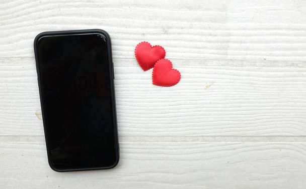 Ein schwarzes Smartphone, Telefon, Handy mit einem Herz-Symbol, umgeben von kleinen roten Herzen isoliert auf einem Holztisch weißen Hintergrund. Minimalistischer Trend.Valentinstag, Dating mobile Anwendung, digital online - Foto, Bild