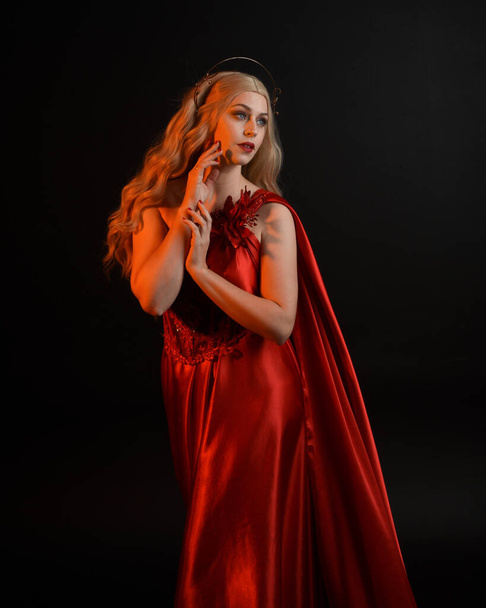 Close up portrait of beautiful blonde model wearing flowing red silk toga gown and crown, dressed as ancient mythological fantasy goddess. Graceful elegant pose isolated on dark studio background. - Photo, Image
