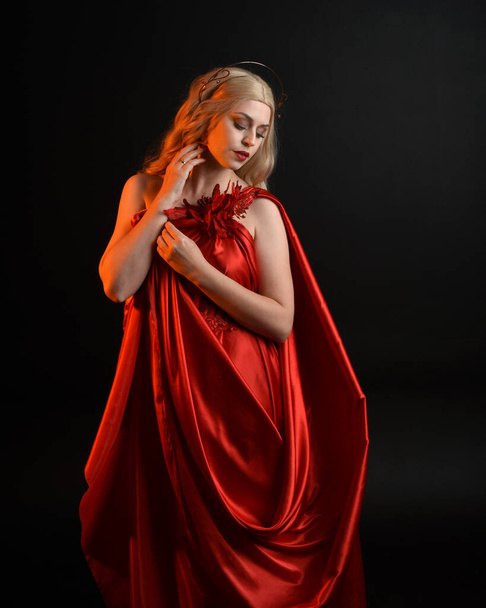 Close-up portret van mooi blond model met vloeiende rode zijden toga jurk en kroon, verkleed als oude mythologische fantasiegodin. Graceful elegante pose geïsoleerd op donkere studio achtergrond. - Foto, afbeelding