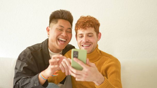 Mutlu çok ırklı eşcinsel çift evdeki cep telefonundan bir şeye bakıyor. - Fotoğraf, Görsel