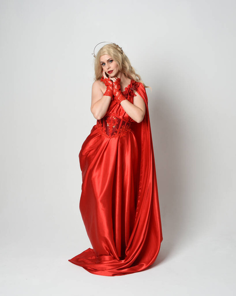 Full length portret van mooi blond model gekleed als oude mythologische fantasie godin in vloeiende rode zijden toga jurk, kroon. elegante dansende pose met vloeiende stof, geïsoleerd op witte studioachtergrond. - Foto, afbeelding