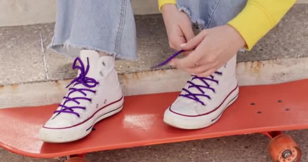 Female teenager ties shoelaces on sneakers standing on skateboard. Girl prepares for skateboarding in jeans and comfortable sporty shoes - Footage, Video