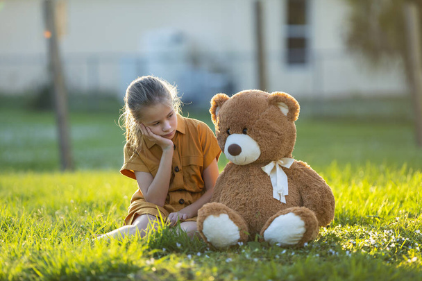 Trauriges einsames Kindermädchen, das mit seinem Teddybärenfreund im sonnigen Hinterhof Zeit im Freien verbringt. Einsamkeit in jungen Jahren. - Foto, Bild
