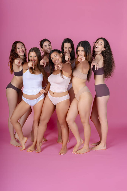 Щасливі різноманітні жінки, що вказують на камеру в дисплеї позитиву тіла, в нижній білизні на рожевому фоні.  - Фото, зображення