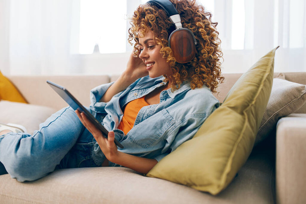Ευτυχισμένη νεαρή γυναίκα απολαμβάνοντας ζεστή χαλάρωση στο σπίτι: Διαβάζοντας, παίζοντας και κουβεντιάζοντας online με ακουστικά στον καναπέ. - Φωτογραφία, εικόνα