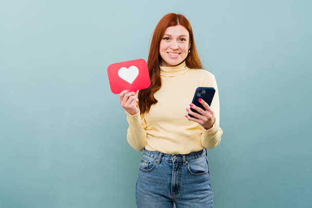 Sosyal medya kullanan güzel kızıl saçlı bir kadının portresi ve mesajlaşırken çevrimiçi bir kalp tuşu göstermek  - Fotoğraf, Görsel