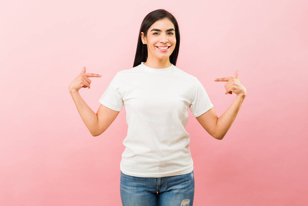 Χαρούμενη νεαρή γυναίκα χαμογελά δείχνοντας τα δάχτυλά της στο λευκό μπλουζάκι της, δείχνοντας χαρούμενη και ενθουσιασμένη. - Φωτογραφία, εικόνα