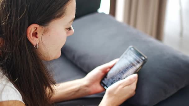 Genç, gülümseyen bir kadın kanepede uzanıyor ve akıllı telefonunda komik bir kitap okuyor. Yüksek kalite 4k görüntü - Video, Çekim