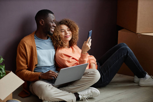Junge Frau zeigt jungen Mann Fotos auf ihrem Smartphone, während er Laptop benutzt, sie sitzen auf dem Boden im Zimmer - Foto, Bild