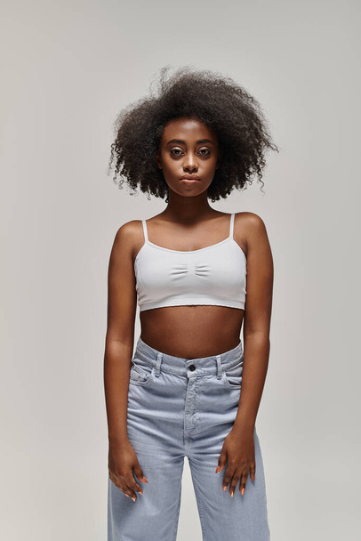 A beautiful African American woman with curly hair posing in a stylish white crop top and jeans in a studio setting. - Photo, Image