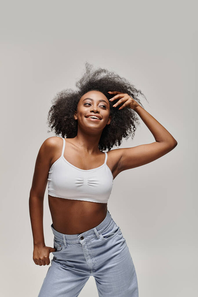 Krásná mladá Afroameričanka s kudrnatými vlasy v bílém svršku a džínách, vyzařující sebevědomí a styl. - Fotografie, Obrázek