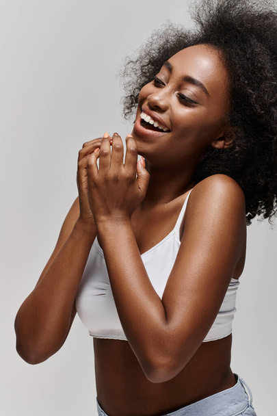 Una hermosa joven afroamericana con el pelo rizado vistiendo un top blanco, sonriendo brillantemente y aplaudiendo. - Foto, Imagen