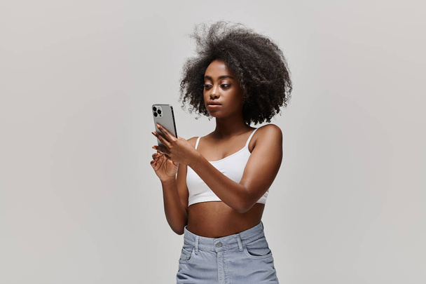 Ohromující Afroameričanka s kudrnatými vlasy drží mobilní telefon, zatímco na sobě bílý top plodin ve studiu nastavení. - Fotografie, Obrázek