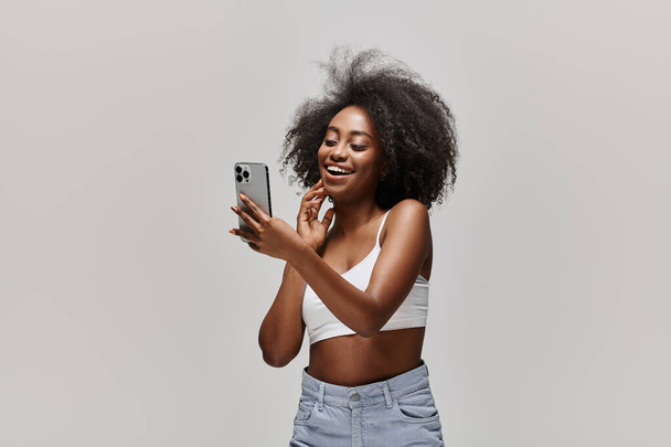 Een stijlvolle Afro-Amerikaanse vrouw in een wit topje, met een mobiele telefoon. - Foto, afbeelding