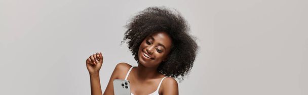 Eine schöne junge Afroamerikanerin mit lockigem Haar hält ein Handy in der Hand, während sie ein weißes Tank-Top trägt. - Foto, Bild