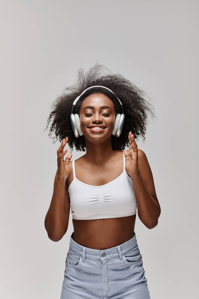 Eine schöne Afroamerikanerin mit lockigem Haar steht vor einer grauen Kulisse und trägt Kopfhörer. - Foto, Bild