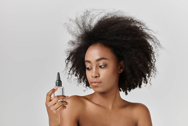 Nuori afroamerikkalainen nainen, jolla on kiharat hiukset, pitää hiuspulloa, parantaa hänen kauneuttaan ja korostaa kehon hoitoa.. - Valokuva, kuva