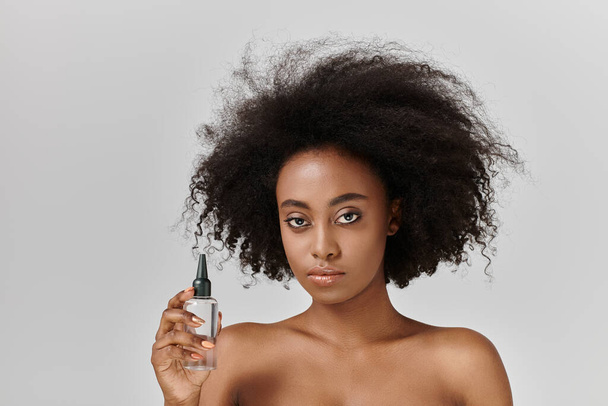 Μια όμορφη νεαρή Αφροαμερικανή γυναίκα με σγουρά μαλλιά που κρατάει ένα μπουκάλι προϊόν για τα μαλλιά, προωθώντας την έννοια της φροντίδας του δέρματος και της αυτοαγάπης. - Φωτογραφία, εικόνα