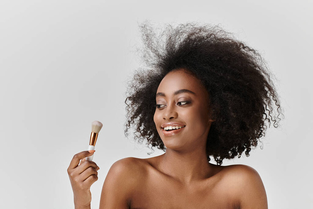 Egy fiatal, göndör hajú afro-amerikai nő ecsetet tart, a bőrápolásra és a szépségápolásra összpontosítva egy stúdióban.. - Fotó, kép