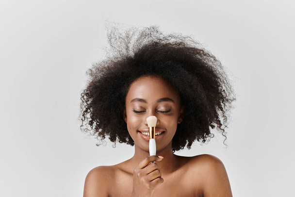 A beautiful young African American woman with curly hair holding a brush near her mouth in a skincare concept studio setting. - Photo, Image