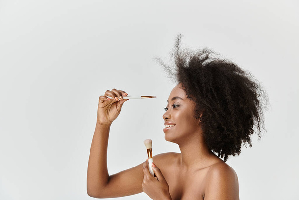 Μια νεαρή Αφρο-Αμερικανίδα γυναίκα με σγουρά μαλλιά απλώνει μια βούρτσα στο πρόσωπό της ως μέρος μιας ρουτίνας φροντίδας του δέρματος.. - Φωτογραφία, εικόνα